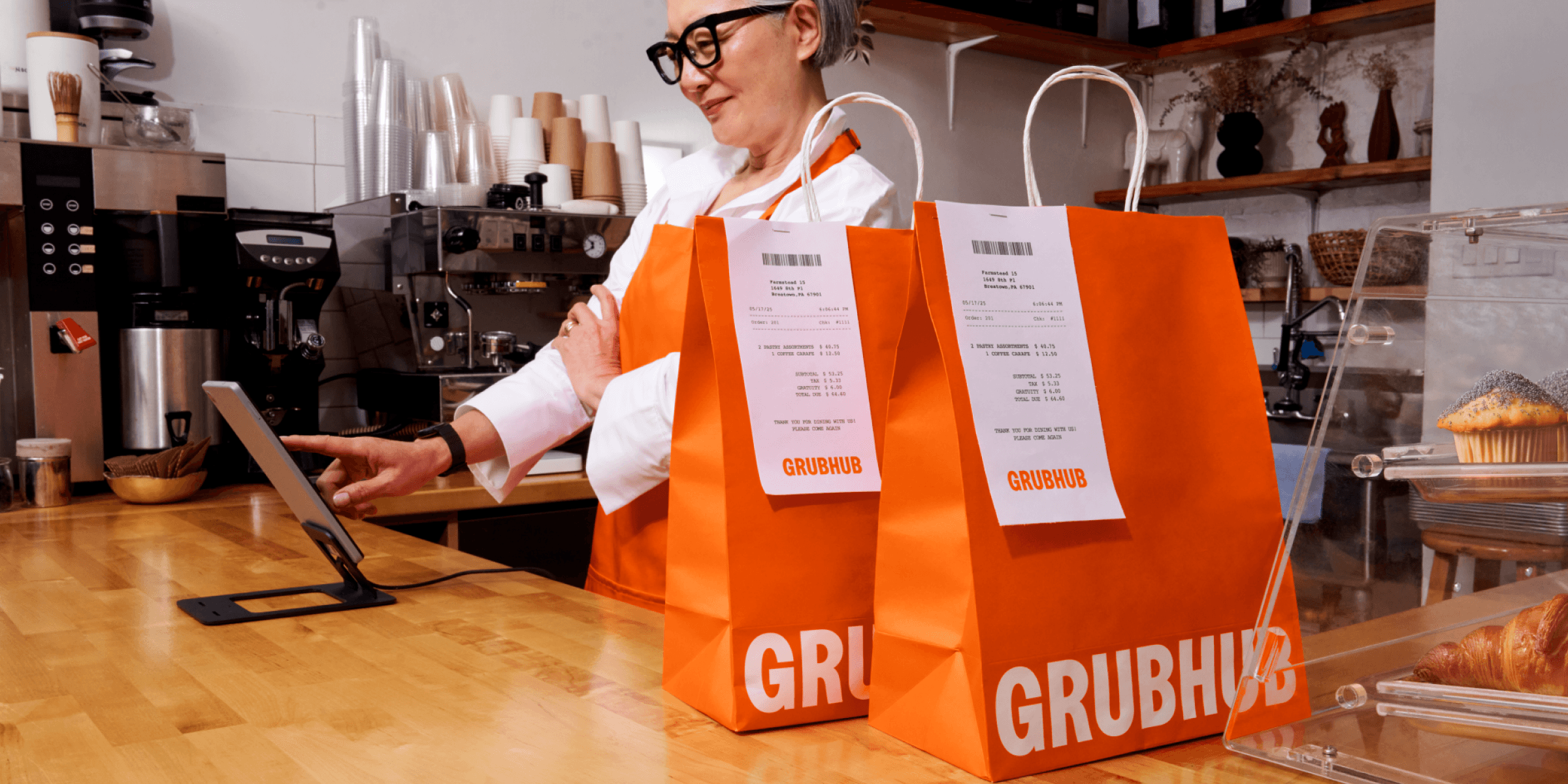 two orange Grubhub bags on a counter, a woman in the background using a tablet