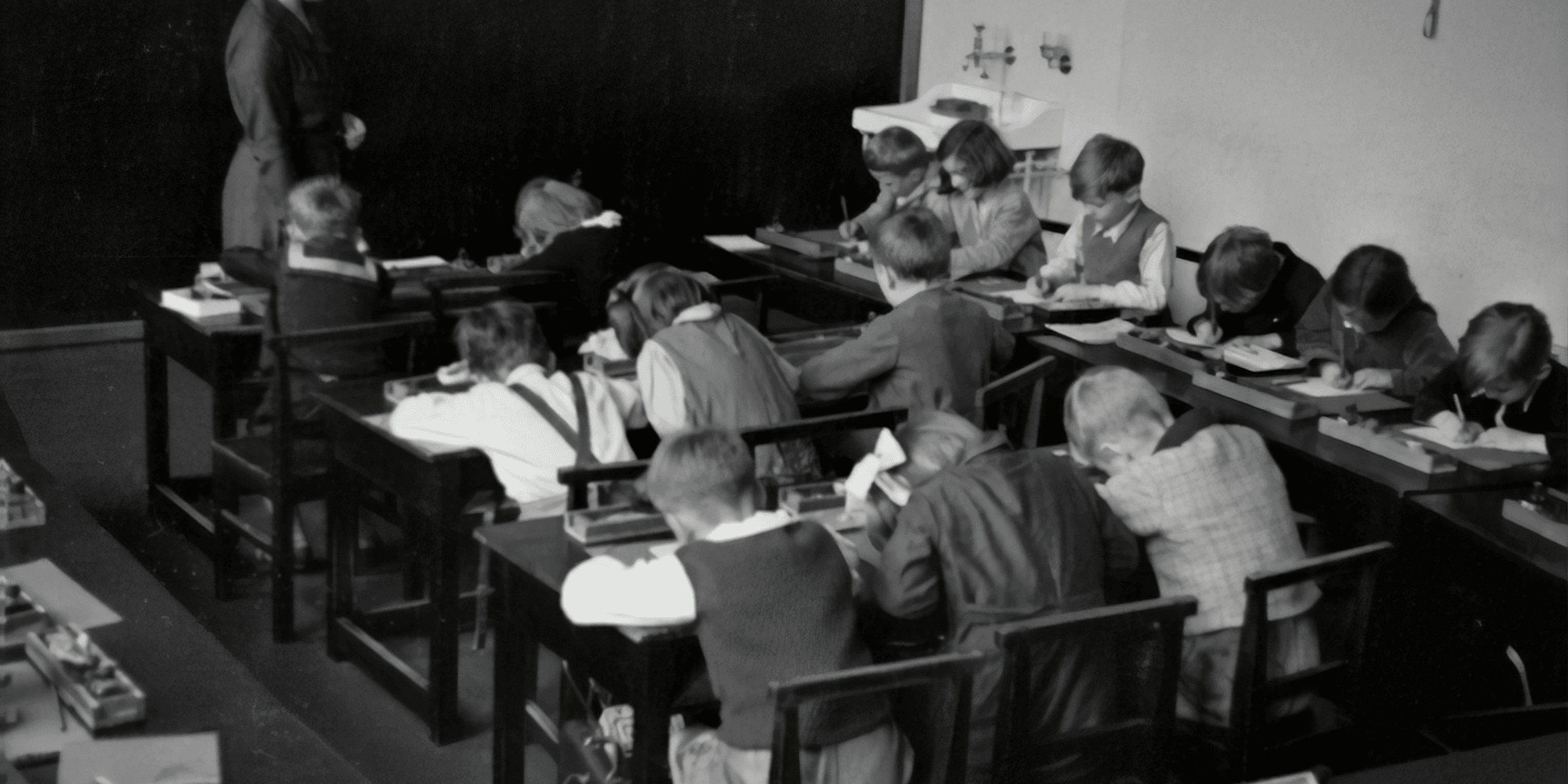 grayscale photography of teacher standing near chalkboard and children sitting on chairs.