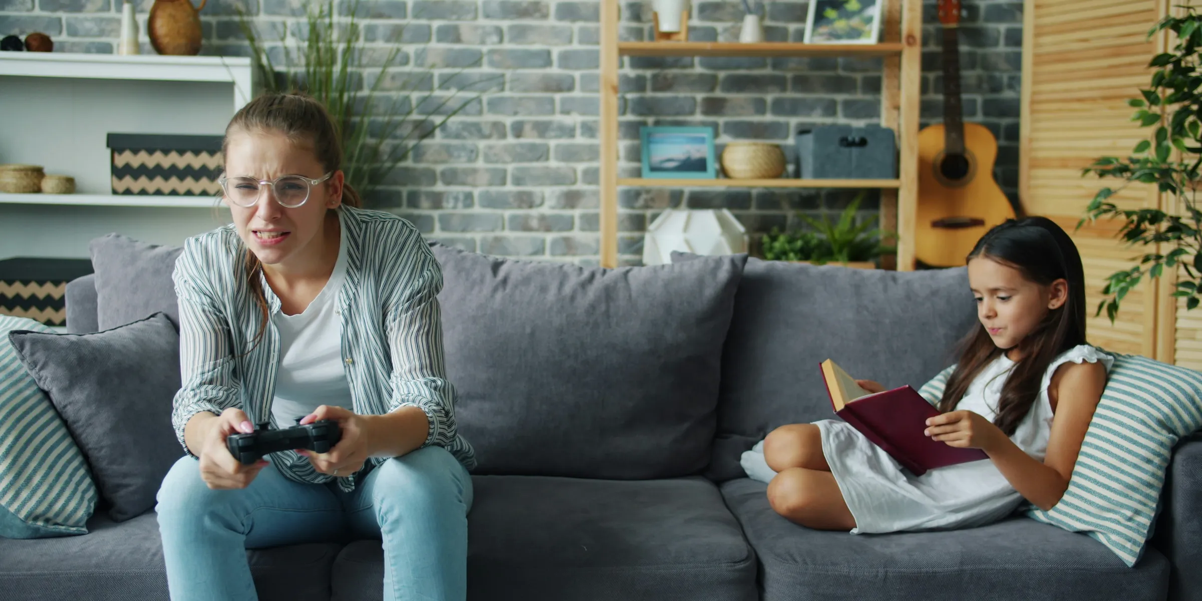 Woman plays video game while girl reads book on couch.