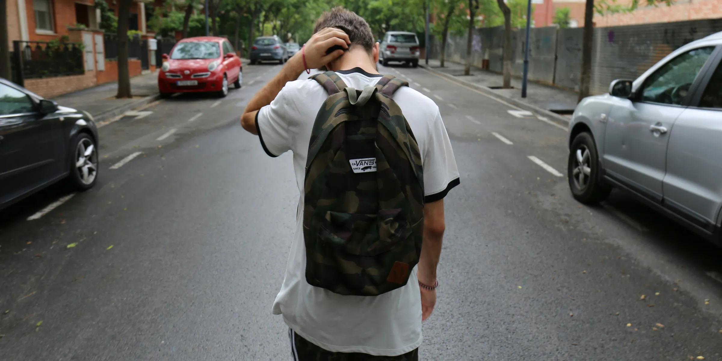 boy wearing white shirt and black shorts carrying backpack standing on black concrete road between vehicles and trees during daytime
