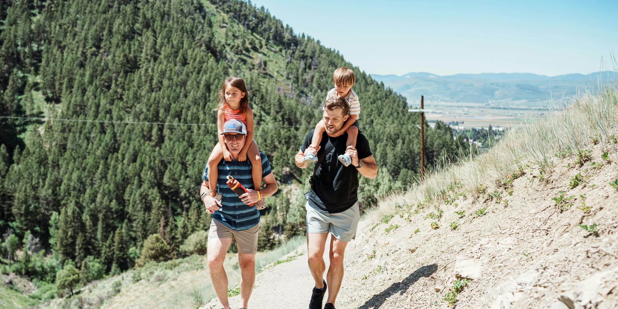 Two fathers carrying children on shoulders on a mountain path.