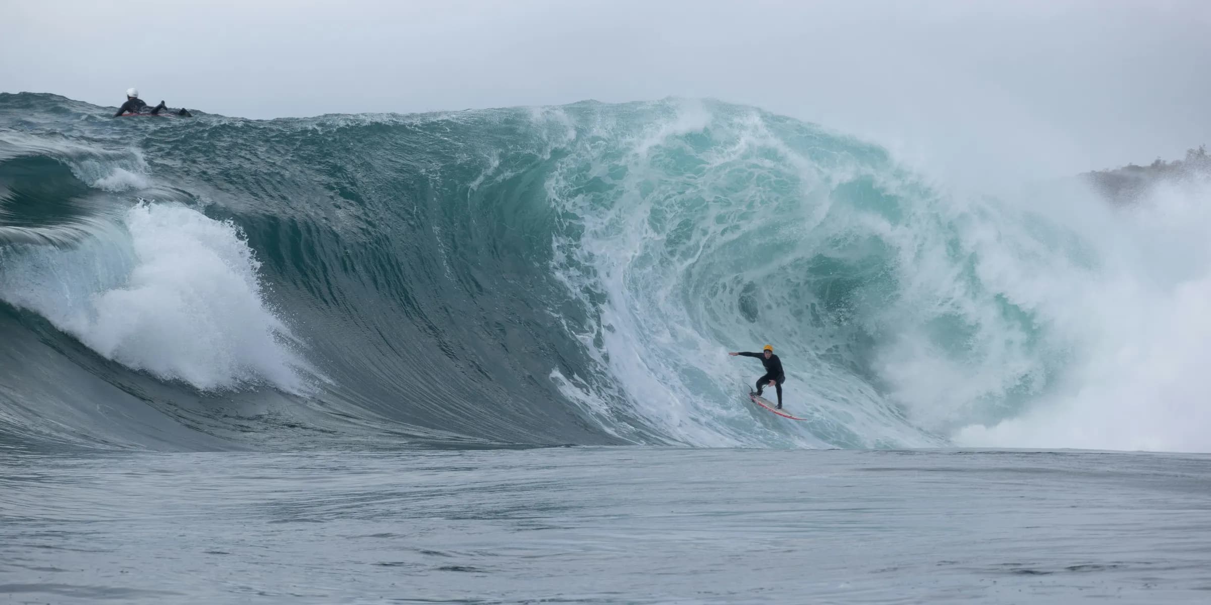 Surfer riding a massive wave under a cloudy sky.