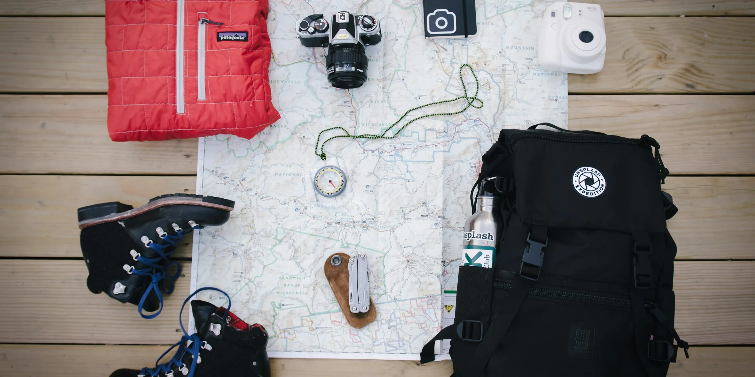 black hiking backpack near white Fujifilm instax mini camera near black leather boots, red half-zip jacket, gray pocket watch on white map