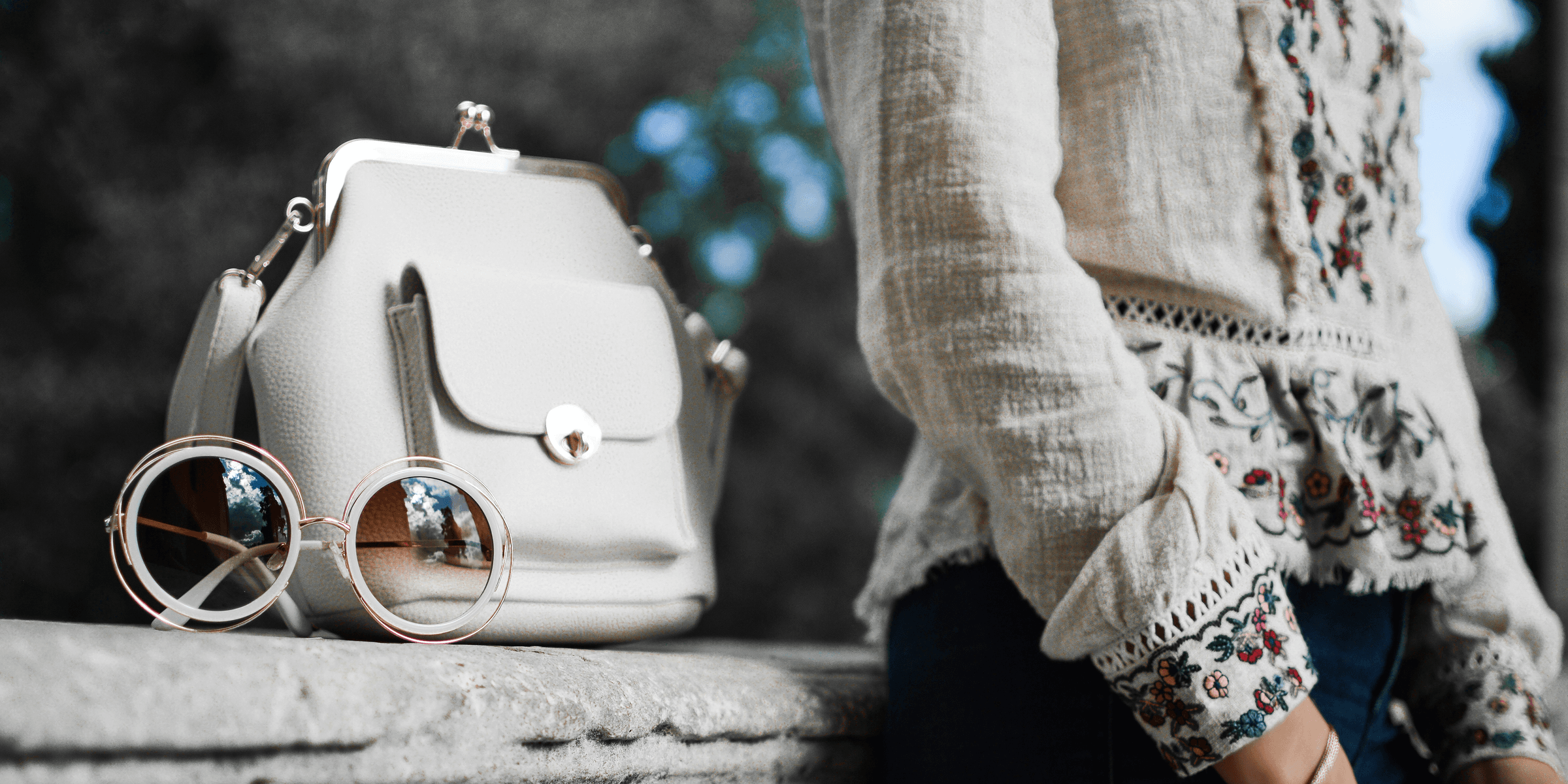 woman wearing beige and red floral top leaning on gray concrete slab with white leather bag ontop.
