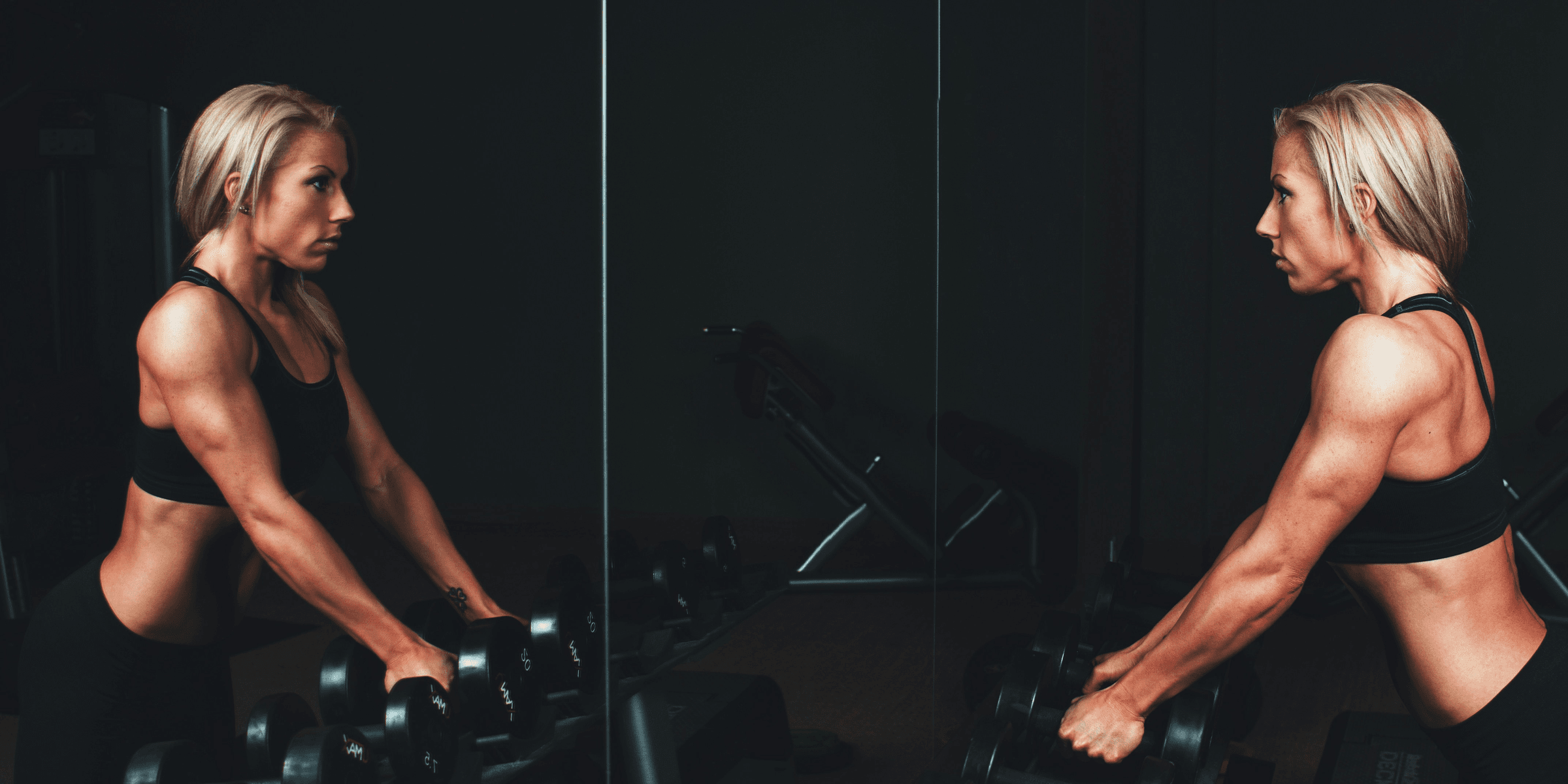woman wearing black top top holding black dumbbells standing in front of mirror.