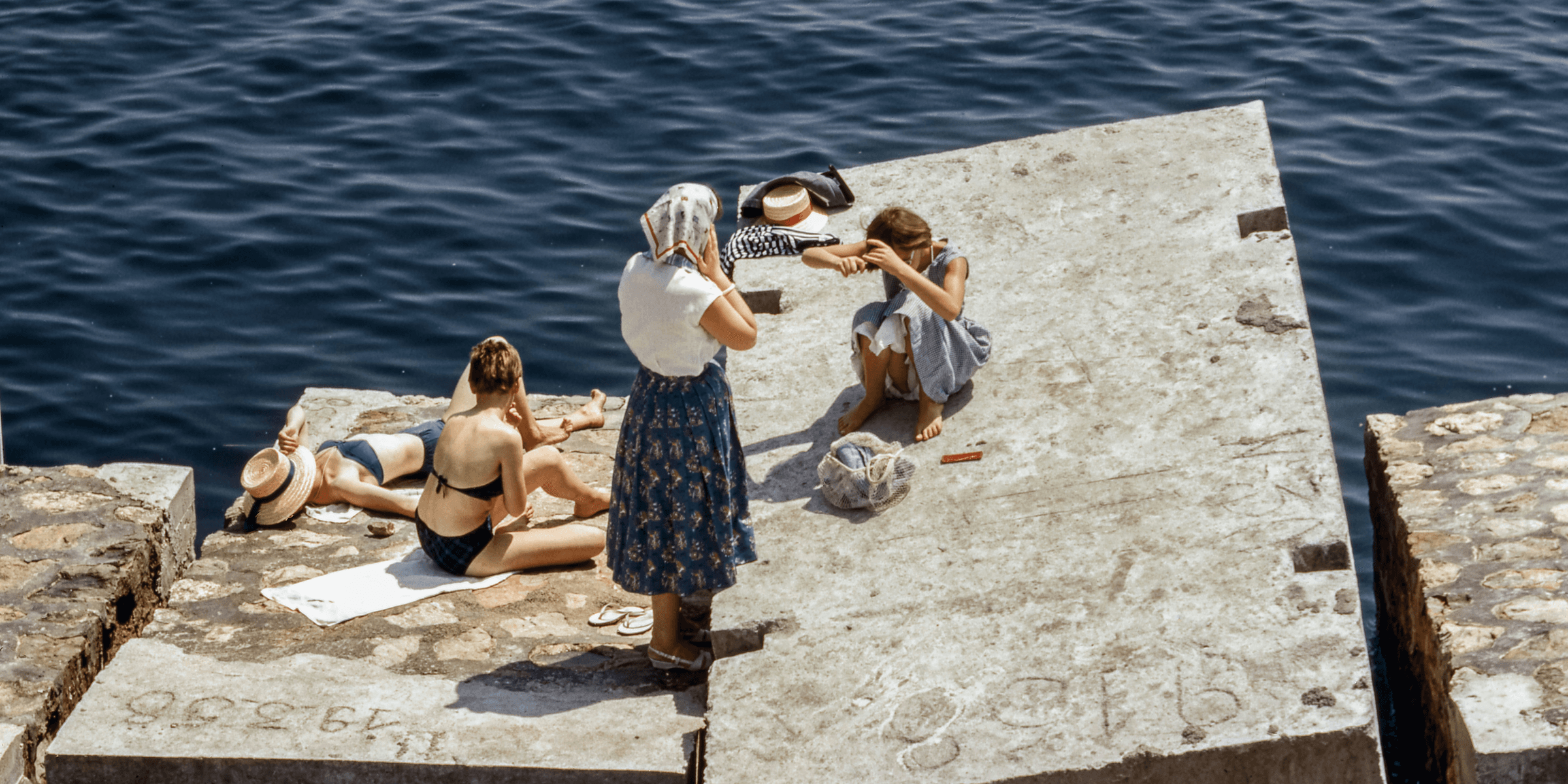 Women in swimsuits and vintage clothing sunbathing on cement cubes by the sea.