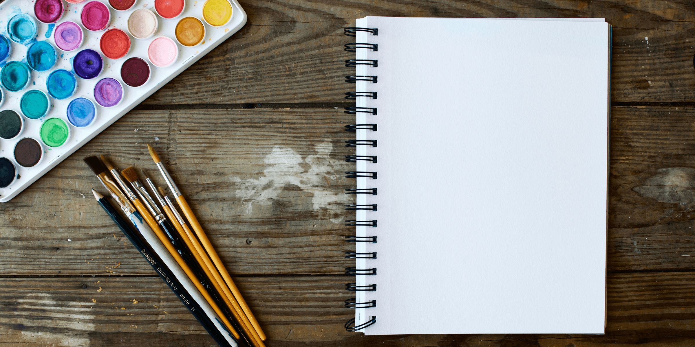 paint brushes next to drawing book and water color palette.