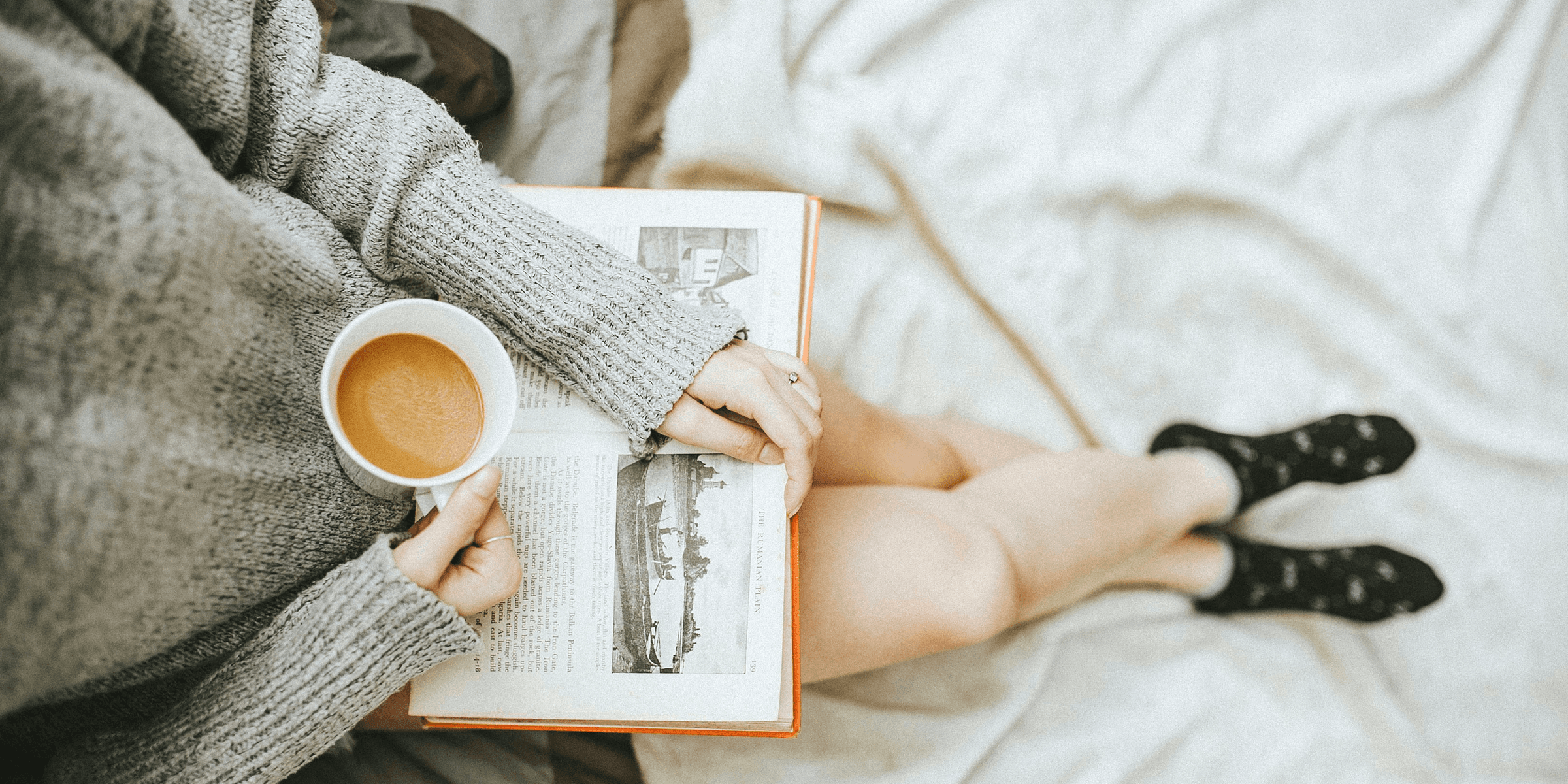 woman holding a cup of coffee at right hand and reading book on her lap while holding it open with her left hand in a well-lit room.