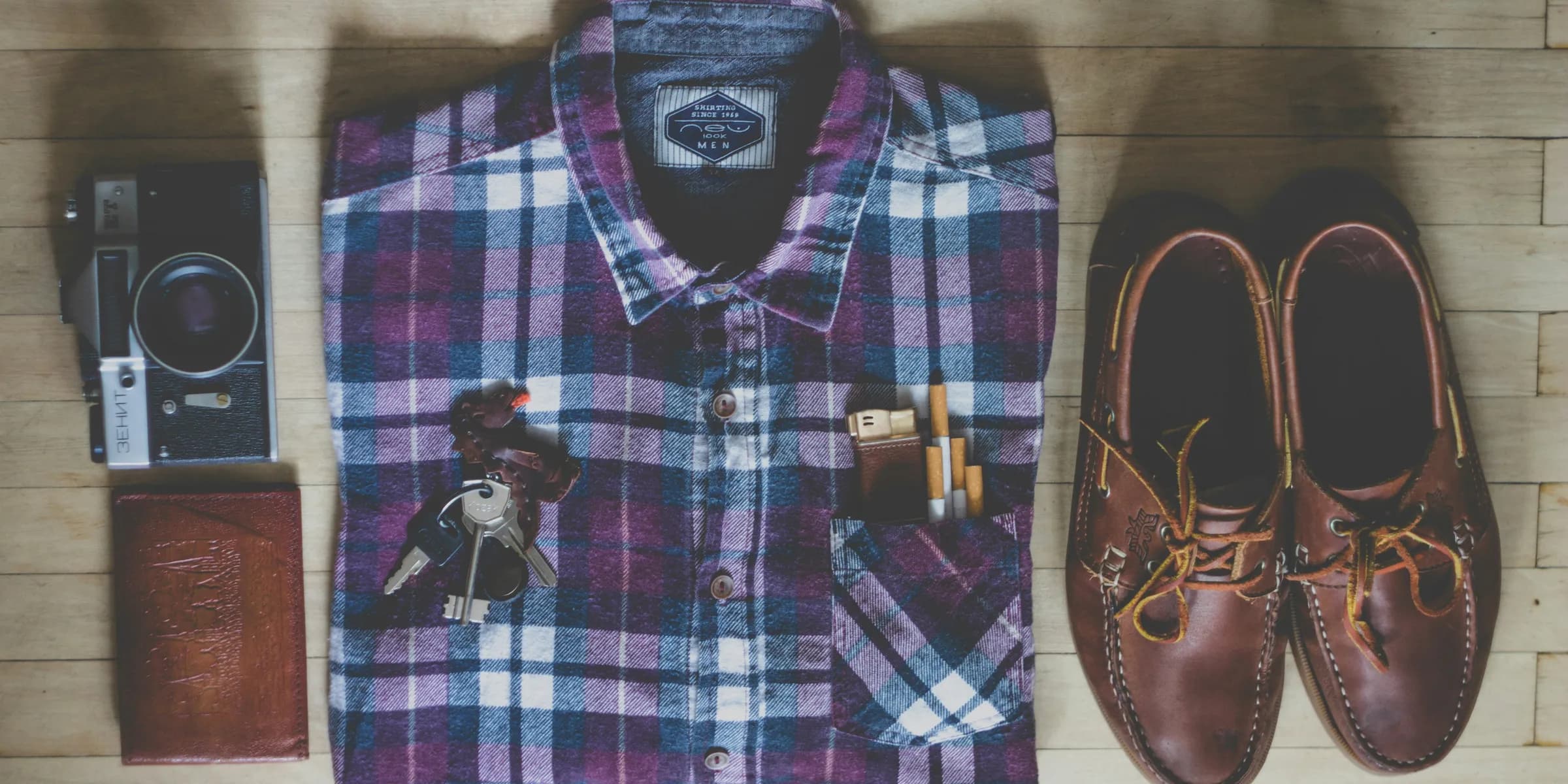 flat lay photography of button-up shirt, camera, keys, cigarettes, and pair of brown leather boat shoes