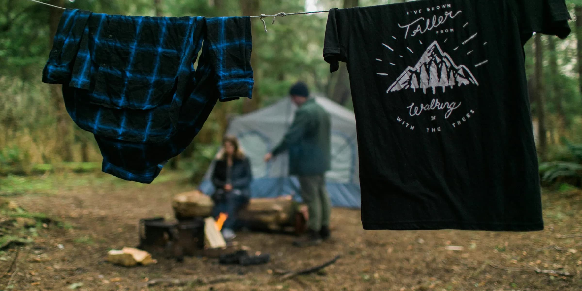 two shirts hanging near woman and man beside blue tent under tall trees