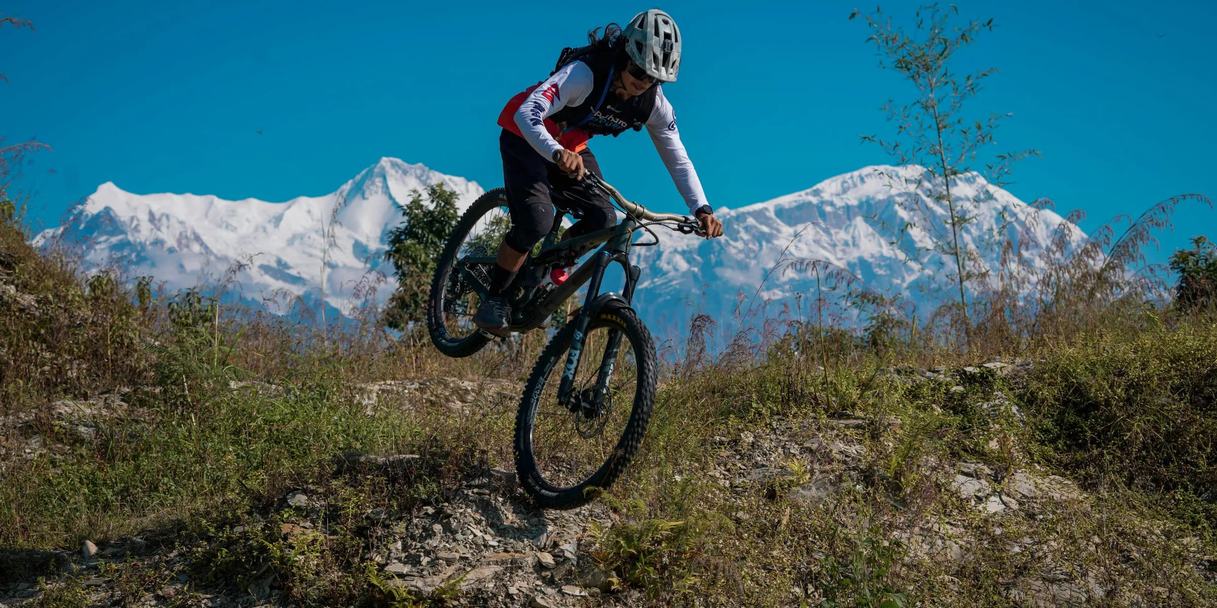 Mountain biker jumps over a dirt mound with snow-capped mountains.
