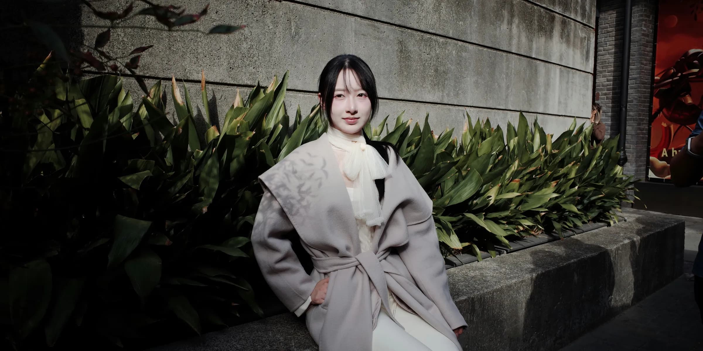 Young woman in stylish coat sitting outdoors near plants.
